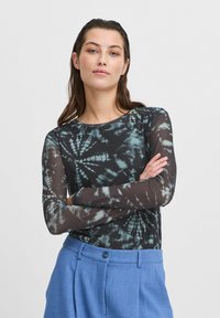 Schwarzes Langarmshirt mit blau-grünem Batikmuster; durchsichtiges Material, runder Ausschnitt, kombiniert mit hellblauen, hochgeschnittenen Hosen.