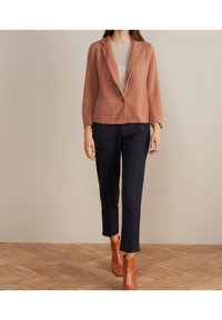 Blazer v rustové barvě s texturou a rukávy po tři čtvrtiny, s jedním knoflíkem, kombinovaný s tmavě modrými šitými kalhotami a hnědými kotníkovými botami.