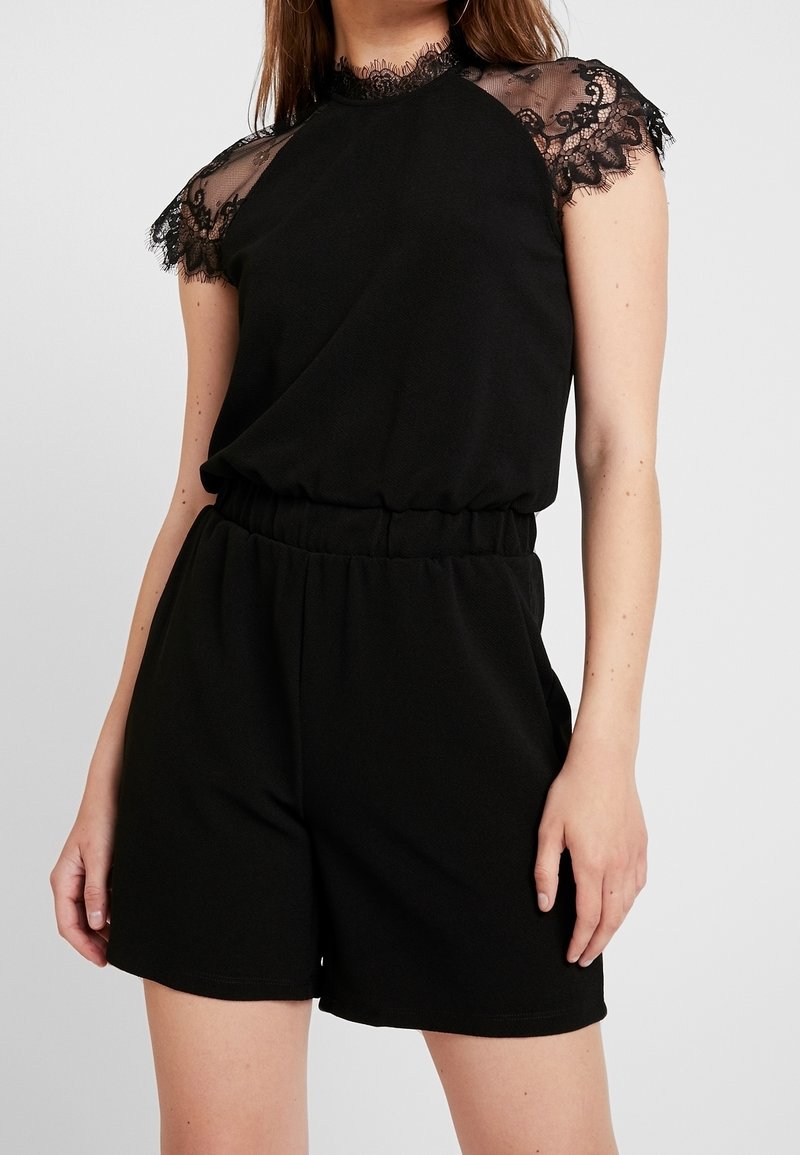 Femme portant une combinaison noire sans manches avec des épaules en dentelle ajourée et une taille haute élastique, debout sur un fond gris.