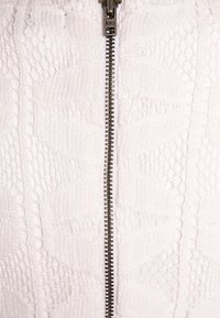 Biała tkanina z teksturowanymi wzorami, z centralnym srebrnym zamkiem błyskawicznym i teksturowanymi akcentami po obu stronach zamka.