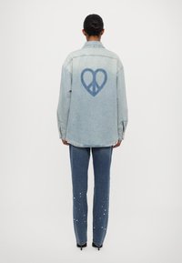 Gaišzilā džinsa jaka ar sirdī ietvertu miera simbolu grafiku aizmugurē, kombinēta ar tumši zilām biksēm, kurām ir izkaisīti dekoratīvie studu elementi.