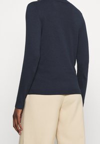 Henkilö, joka on pukeutunut tiukkaan tummansiniseen pitkähiharaiseen puseroon ja löysiin vaaleisiin housuihin, kuvattuna selkäpuolelta yhdellä vaalealla taustalla.