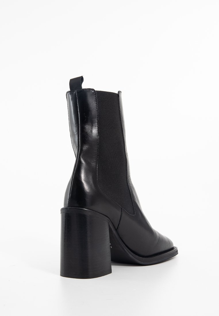 Bottine en cuir noir avec un talon épais, des panneaux latéraux élastiques et une finition lisse. Dispose d'une boucle pour un enfilage facile.