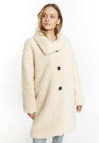 Krémový oversized kabát vyrobený z texturované, sametově hebké látky, s širokým límcem a černými knoflíky vpředu.