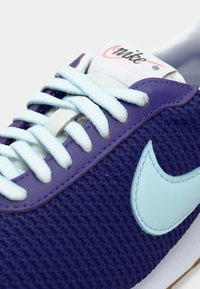 Sapatilha em malha roxa com logo Nike em azul claro, atacadores brancos e etiqueta da língua branca com logo Nike em vermelho e preto.