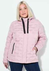 Blond kvinna som bär en ljusrosa quiltad jacka med svarta dragsnören, med händerna i dragkedjefickorna, stående mot en vit bakgrund.