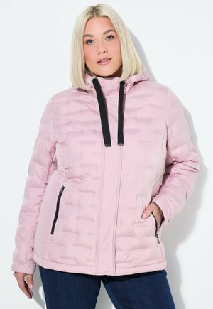 Femme blonde portant une veste matelassée rose clair avec des cordons noirs, les mains dans les poches zippées, se tenant devant un fond blanc.