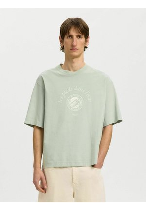 Fiatal férfi áll, egyik kezével a zsebében, világoszöld bő pólót visel, rajta francia szöveg, bézs nadrággal, fehér háttér előtt.