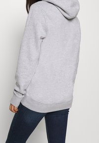 Felpa grigia con cappuccio regolabile, polsini e orlo a costine, tessuto morbido e vestibilità comoda, abbinata a jeans blu scuro.