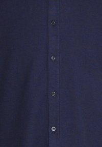 Tela azul marino con cuatro botones oscuros cosidos verticalmente a lo largo de una tapeta de camisa con costuras visibles.