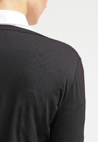 Persona vista da dietro che indossa un blazer nero sopra una camicia bianca con colletto, su uno sfondo grigio chiaro uniforme.