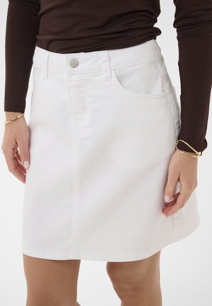 Žena nosi bielu rifľovú sukňu s prednými vreckami a gombíkom, v kombinácii s tmavohnedým dlhým rukávom a zlatými náramkami.