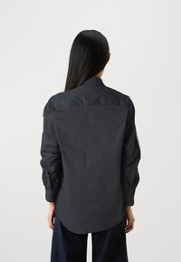Black long-sleeve shirt with a collared neck and buttoned cuffs. Features textured fabric and a curved hem, worn over dark trousers.