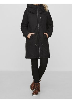 Kobieta ubrana w długi czarny zimowy płaszcz z kapturem i przednimi kieszeniami, czarne spodnie i brązowe sznurowane buty, stojąca z rękami w kieszeniach.