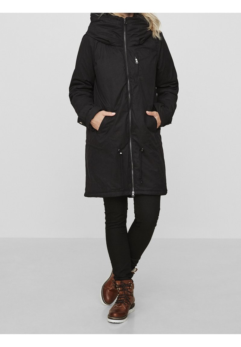 Femme portant un long manteau d'hiver noir à capuche avec poches avant, pantalon noir et bottes marron à lacets, debout les mains dans les poches.
