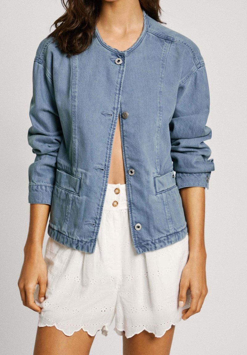 Chaqueta de mezclilla azul clara con cuello redondo, cierre de botones y dos bolsillos frontales. Combinada con pantalones cortos blancos con borde festoneado que presentan detalles de ojales.