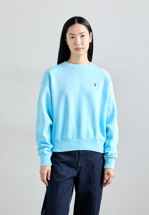 Lys blå hettegenser laget av bomull, med rund hals og ribbede ermer. Liten logo detalj på venstre bryst. Kombinert med mørke jeans.