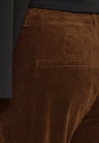 Détail de la poche d’un pantalon en velours côtelé marron sur une personne portant un haut noir, montrant la texture et les coutures du tissu.