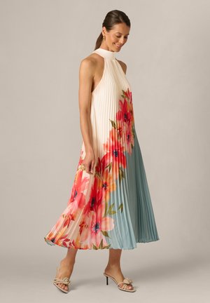 Uśmiechnięta kobieta, ubrana w bezrękawnikową plisowaną sukienkę midi z dużym czerwono-różowym wzorem kwiatowym i jasnoniebieskim blokiem kolorów, stojąca na neutralnym tle.