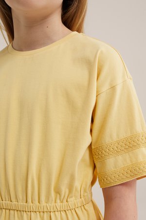 Femeie purtând o rochie galbenă cu mâneci scurte, talie elastică și benzi decorative din dantelă pe mâneci, pe un fundal simplu.