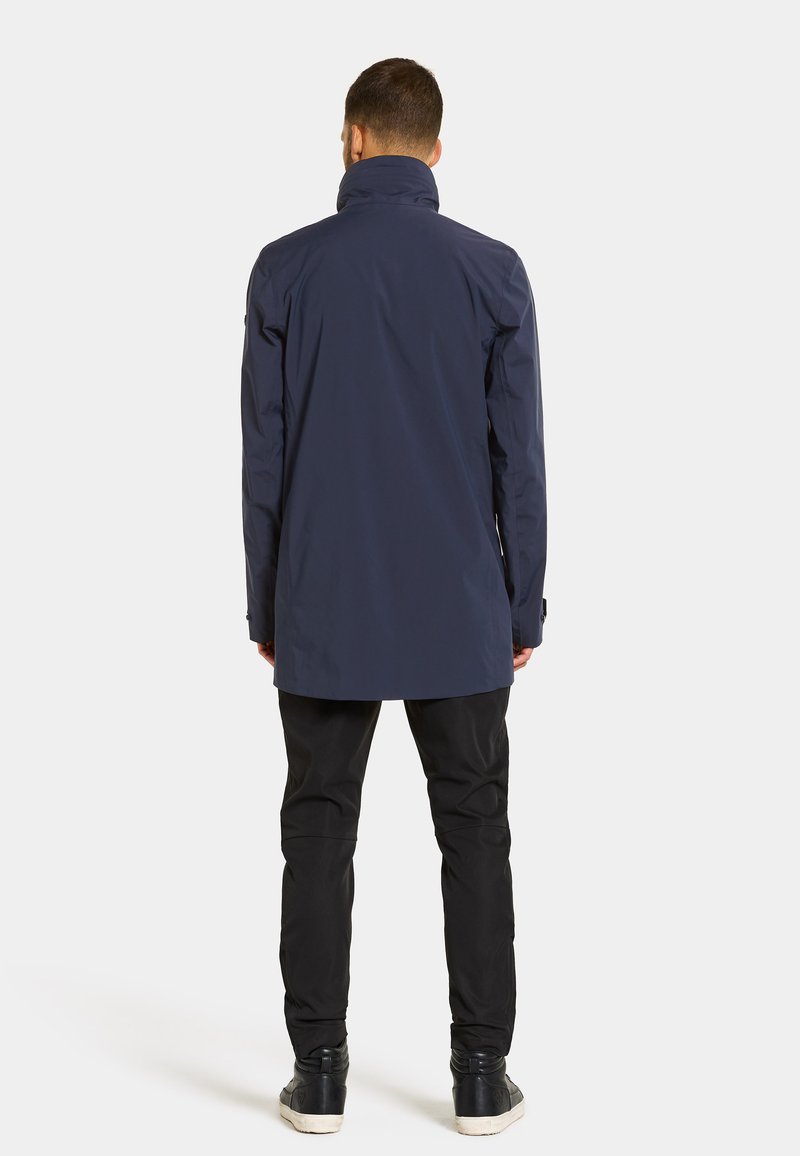 Marineblaue wasserdichte Jacke mit hohem Kragen, langen Ärmeln und einer leicht taillierten Passform, kombiniert mit schwarzen Hosen und dunklen Sneakers.