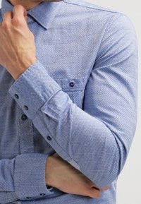 Muž upravující límec světle modré košile s dlouhými rukávy, vzorované, s knoflíkovými manžetami a náprsní kapsou.