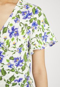 Robe portefeuille florale avec un fond blanc orné de fleurs violettes et de feuilles vertes. Manches courtes à bords volantés. Texture de tissu lisse.