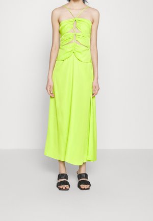 Mujer vestida con un vestido midi sin mangas de color verde lima con recortes fruncidos y sandalias negras de punta abierta, de pie contra un fondo liso.