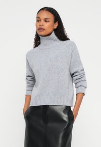 Kvinne iført en lys grå høyhalset genser med oppbrettede ermer og et svart skinnskjørt med hendene i lommene mot en ensfarget bakgrunn.