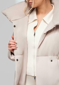 Žena v béžovém prošívaném kabátu přes bílý žebrovaný svetr na zip, drží kabát otevřený, aby odhalila svetr uvnitř.