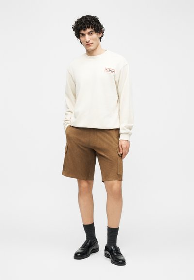 Νεαρός άνδρας που φοράει λευκό μακρυμάνικο πουκάμισο, καφέ cargo σορτς, σκούρες κάλτσες και μαύρα μοκασίνια, στέκεται μπροστά σε ένα απλό φωτεινό φόντο.