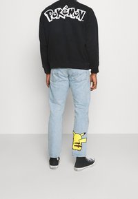 Felpa nera con il logo "Pokémon" sulla schiena; jeans azzurri chiari con una grafica gialla di Pikachu sulla gamba sinistra; sneakers nere.