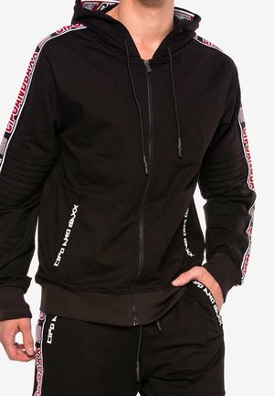 Mann trägt schwarzen Zip-Hoodie und passende Hose mit rot-weißem Markenband entlang der Ärmel und Taschen.