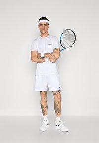 Hvit treningsdrakt bestående av en t-skjorte med kort erme og "BOSS"-logo, matchende shorts, hvite svettebånd og et tenniskølle.