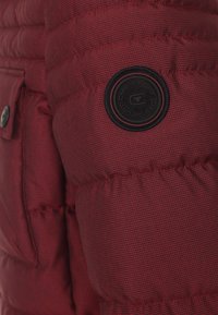 Röd puffjacka med ärm som har kviltat mönster, texturerad tyg och en rund svart logotyp-patch. Inkluderar en synlig tryckknappdetalj.