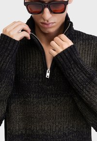 Pull en tricot noir et vert olive avec un col zippé à mi-hauteur. Présente des poignets côtelés et un motif texturé sur l'ensemble.