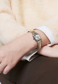 Montre rectangulaire argentée et dorée au poignet, cadran blanc avec accents bleus, bracelet métallique, portée sur un tissu beige et une surface brune.