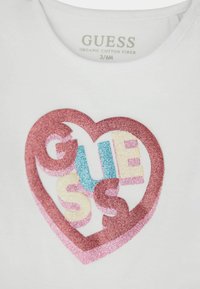 T-shirt bianco in cotone biologico con un cuore glitterato multicolore e il testo "GUESS" in rosa, blu e crema all'interno della forma del cuore.