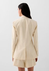 Beige bleiseri, jossa on rakenteelliset hartiat, kaksinkertainen venttiili ja neljän napin hiha. Varmistettu yhdistelemällä vastaaviin shortseihin, sileä kangas.