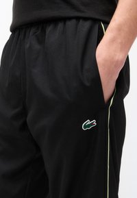 Άτομο που φοράει μαύρο αθλητικό παντελόνι με πράσινες και κίτρινες λωρίδες στο πλάι και λογότυπο κροκοδείλου Lacoste, το χέρι ακουμπισμένο στην τσέπη.