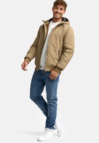 Lysbrun pufferjakke med hætte, to lynlåslommer, parret med blå slim-fit jeans og hvide sneakers. Enkel, funktionel design.