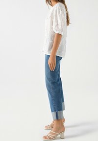 Hvid broderet bluse med pufærmer, parret med blå jeans i denim med rå kant og lyseblå manchet. Strop sandaler i beige.