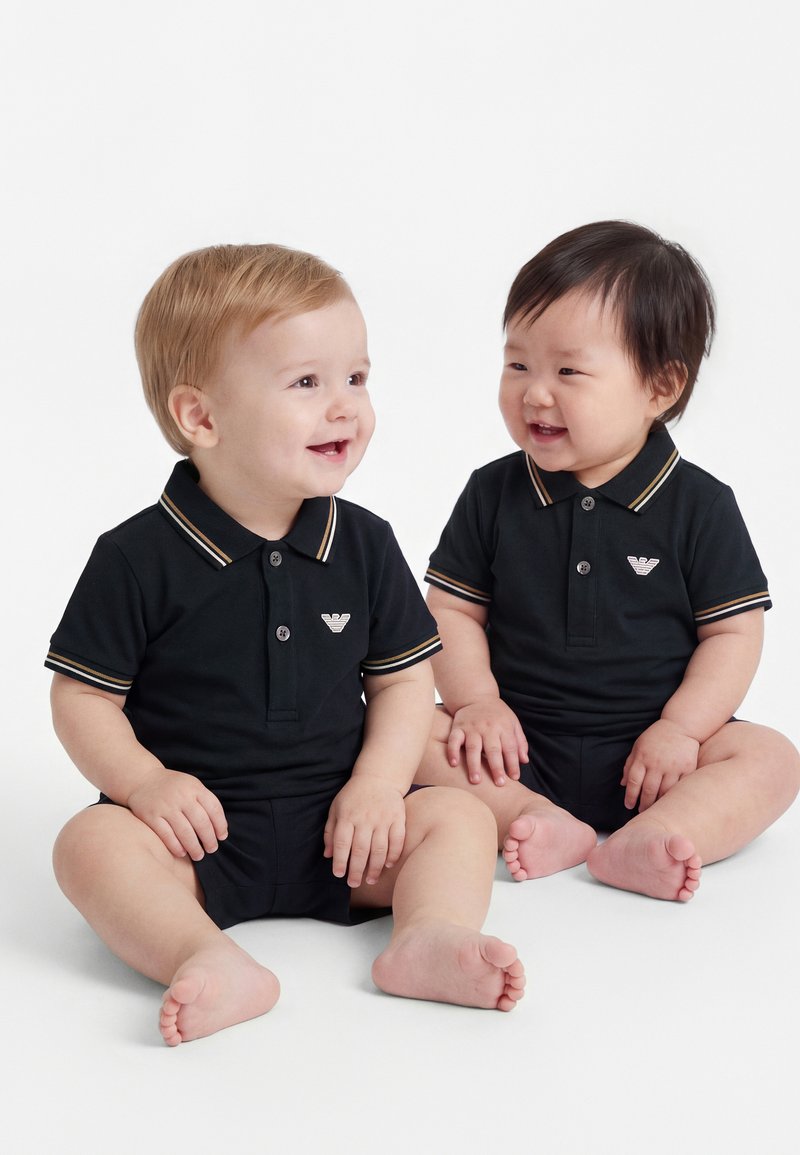 Due neonati sorridenti seduti sul pavimento indossano polo nere abbinate con colletti a righe beige e bianche.