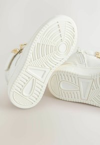 Hvide sneakers med en tekstureret overflade, cirkulært mønster på sålen og hardware i guldfarve nær anklen.