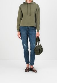 Sweat à capuche vert olive avec une poche avant, associé à un jean bleu déchiré. Chaussures plates rayées noires et marron. Tenant un sac vert foncé.