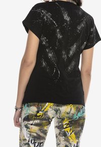 Camiseta negra de algodón con diseño de salpicaduras de pintura blanca, de manga corta y cuello redondo, combinada con pantalones coloridos, estampados y desgastados.