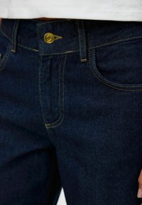 Jeans en denim bleu foncé avec des coutures jaunes contrastantes, un fermoir à bouton doré et des poches avant. Vue rapprochée de la taille.
