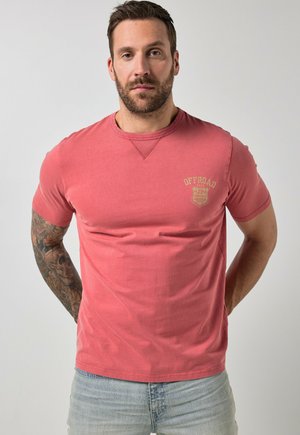 Mann mit kurzem dunklem Haar und Bart, der ein verblasstes rotes Offroad-Grafik-T-Shirt und hellblaue Jeans trägt und mit den Händen hinter dem Rücken steht.