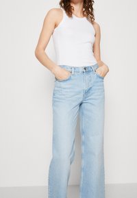 Bílý žebrovaný top s ramínky spárovaný se světle modrými širokými džínami. Džíny mají dvě přední kapsy a zapínání na knoflík v pase.