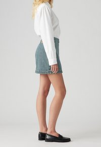 Minifalda de mezclilla en verde azulado desgastado con un dobladillo deshilachado. Combinada con una camisa blanca de manga larga y unos mocasines negros.